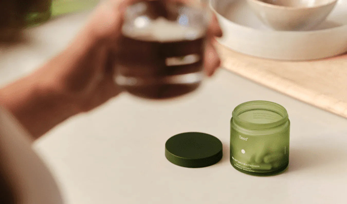 Person holding a glass container with a green lid on a light surface.